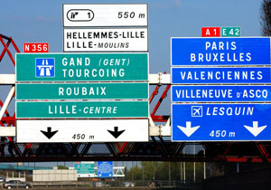 Green and blue directional signs suspended above a road, indicating routes to key destinations such as Lille, Paris, Brussels, Lesquin airport, and more.