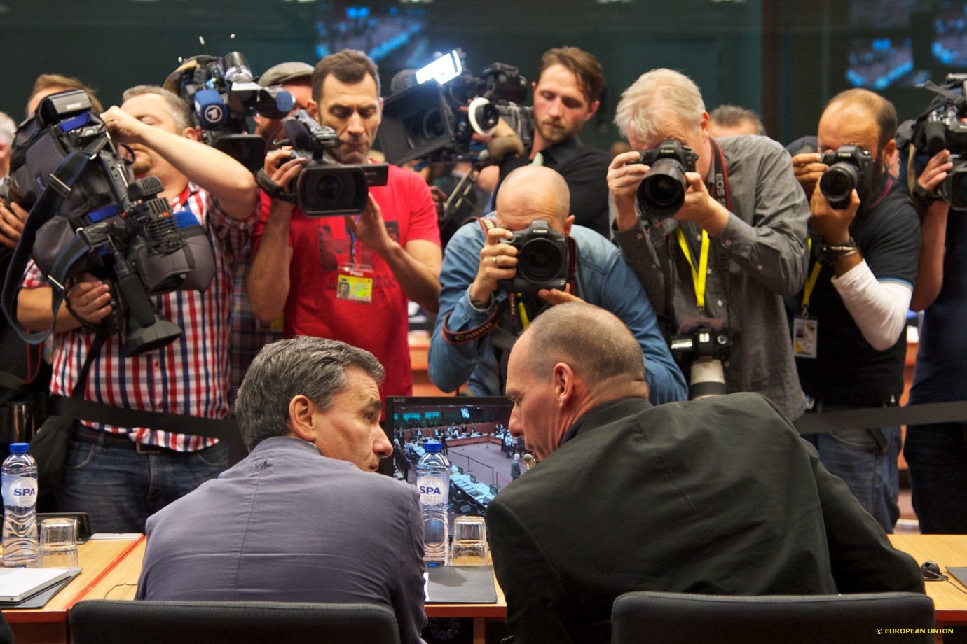 Two men sitting at a table and talking together. A large group of press photographers in front of them.