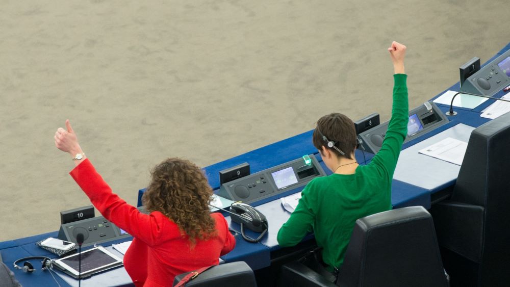 Two women in the European Parliament raising their hands with a thumbs up. Sitting in their seats with backs to camera.