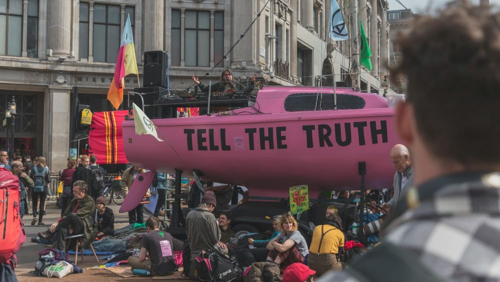 People in a rally against global warming, with a large, pink sail boat in the middle of the road. Text on the boat reads: tell the truth.