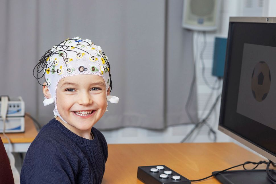 A smiling child, wearing a cap for research purposes, sitting at a desk.